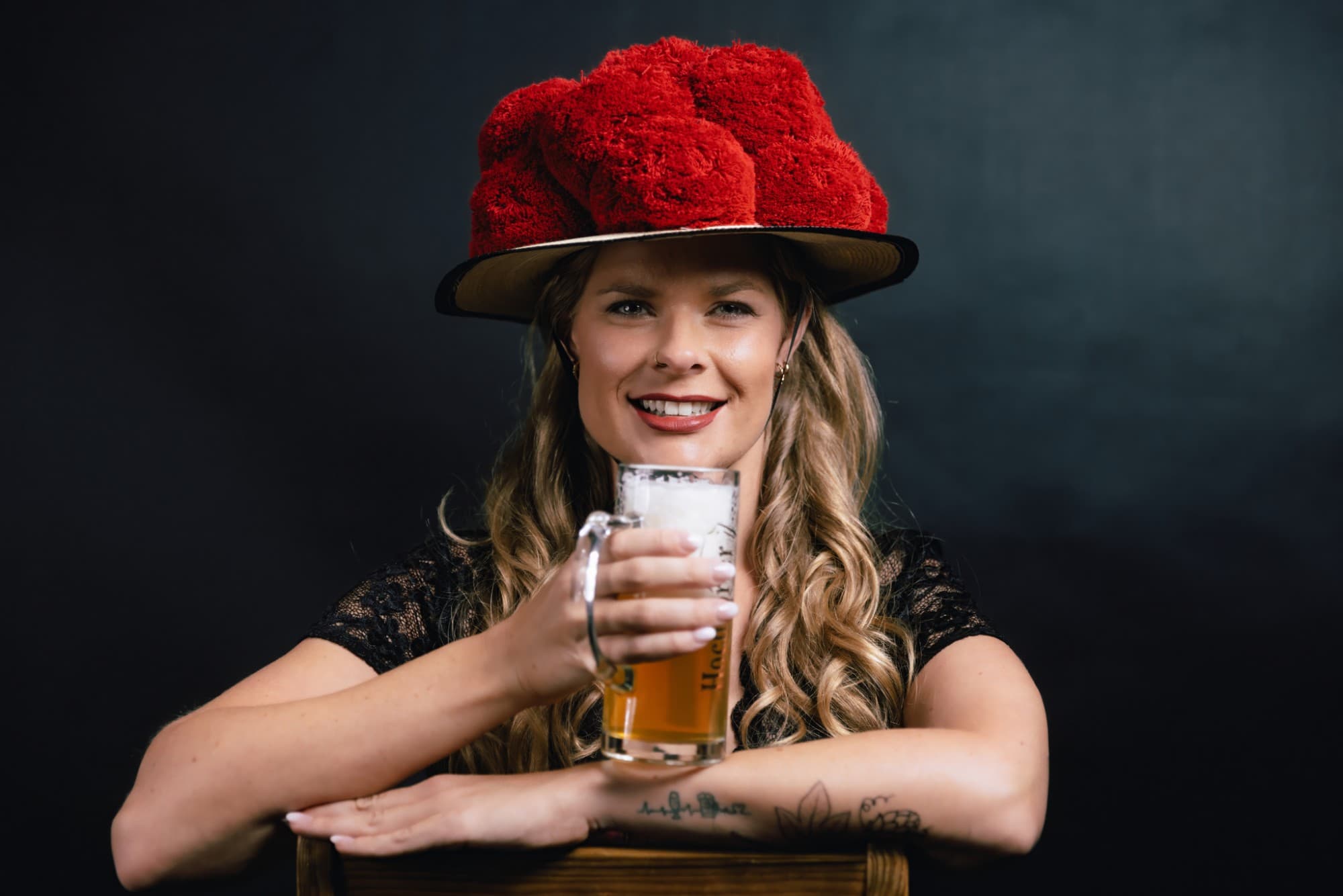 Mary lehnt mit Bollenhut über die Lehne eines Holzstuhls und hält einen Krug helles Bier in die Kamera.