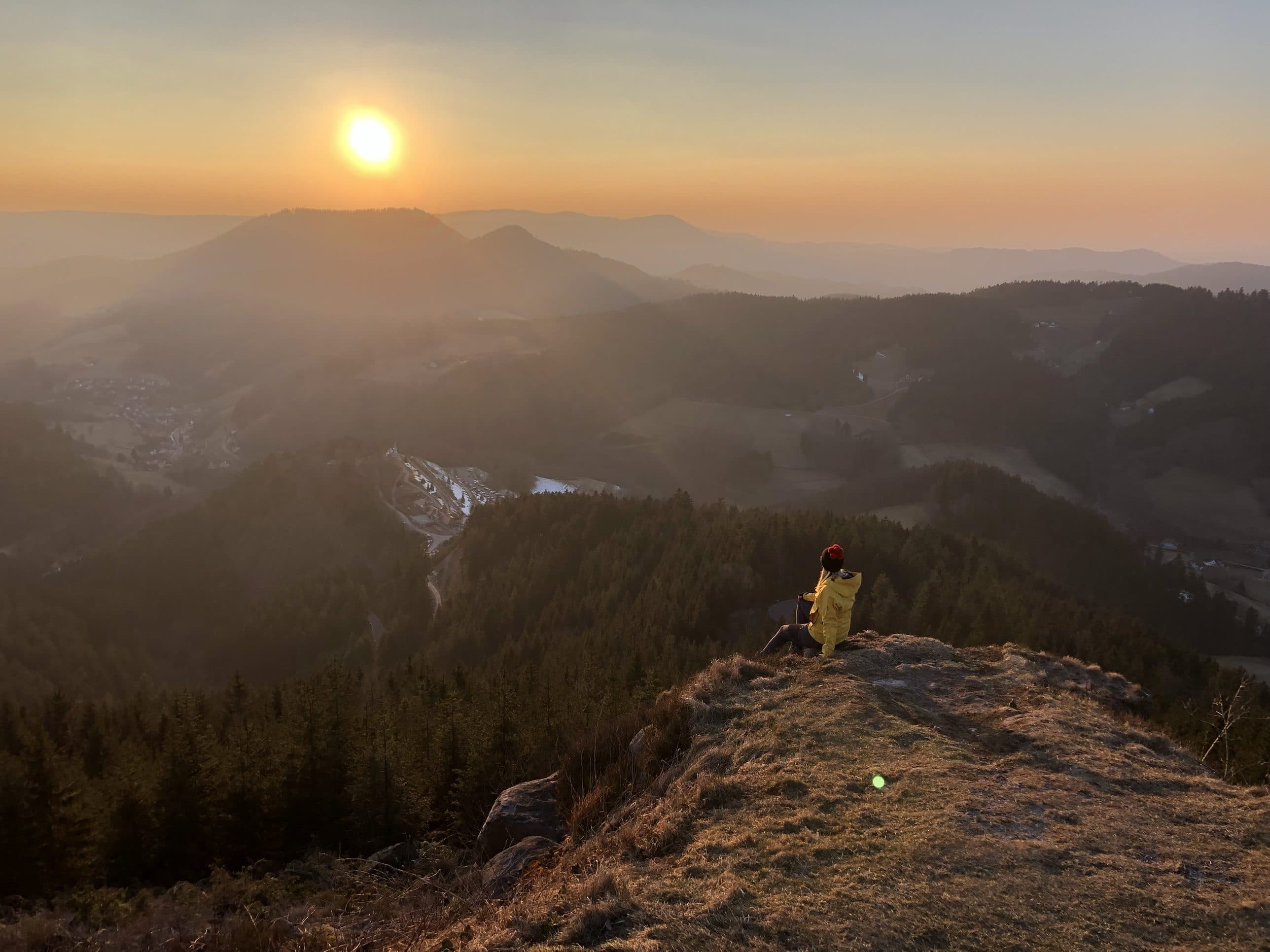 Mary sitzt mit Mütze und gelber Jacke auf einer Bergkuppe und schaut über bewaldete Hügel im Dunst in den Sonnenuntergang.